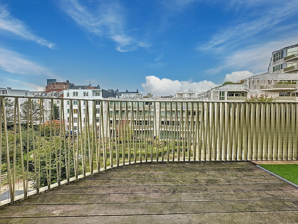 Balcon urbain en bois avec vue sur bâtiment moderne, ciel bleu et verdure apaisante.