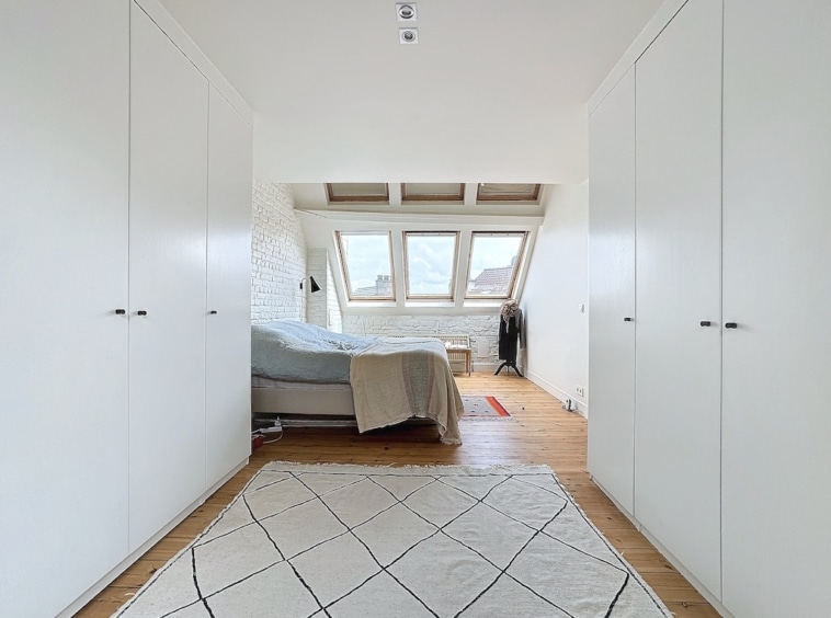 Chambre minimaliste lumineuse avec lit neutre, grandes fenêtres, tapis géométrique et rangements intégrés.