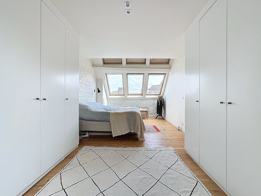 Chambre minimaliste lumineuse avec lit neutre, grandes fenêtres, tapis géométrique et rangements intégrés.