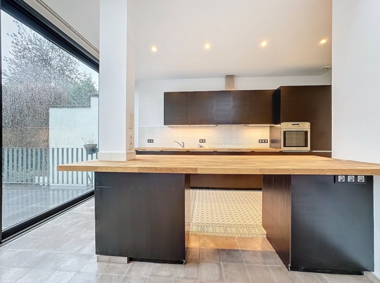 Cuisine moderne lumineuse avec grandes fenêtres, îlot en bois, armoires foncées et vue verdoyante.
