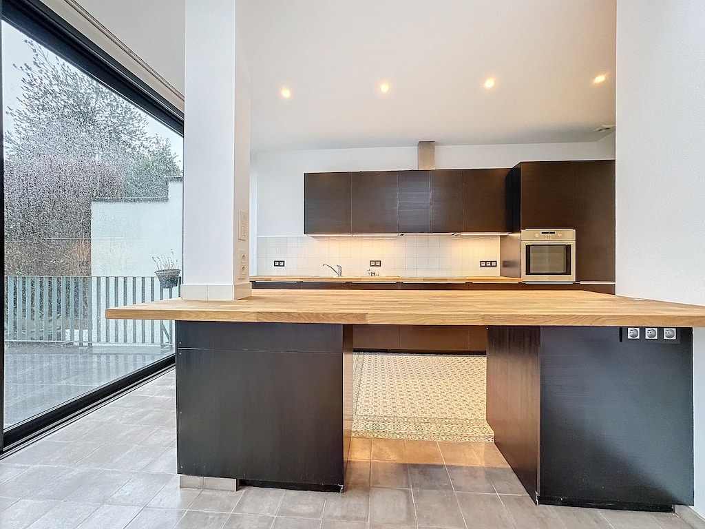 Cuisine moderne lumineuse avec grandes fenêtres, îlot en bois, armoires foncées et vue verdoyante.