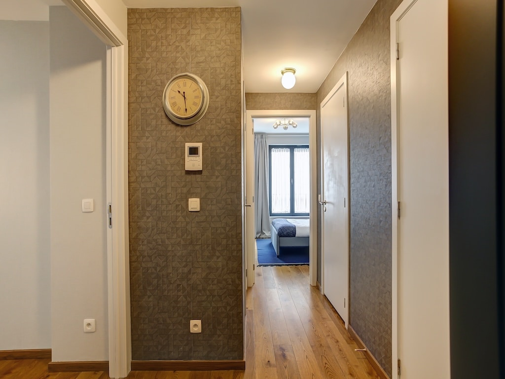 Couloir moderne lumineux avec murs fleuris, sols en bois, grandes fenêtres, portes blanches élégantes.