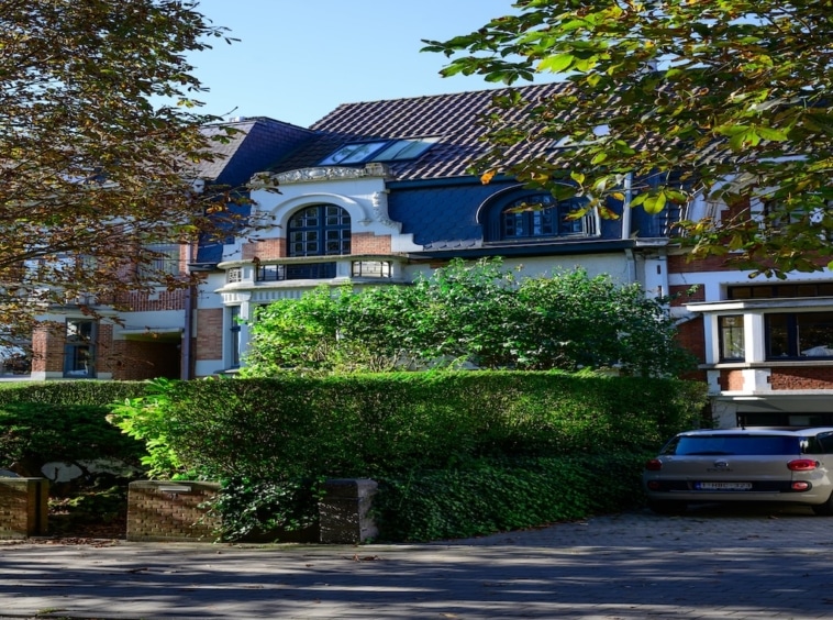 Maison moderne avec toit bleu, jardin paysager, balcon, allée pavée et vue dégagée.