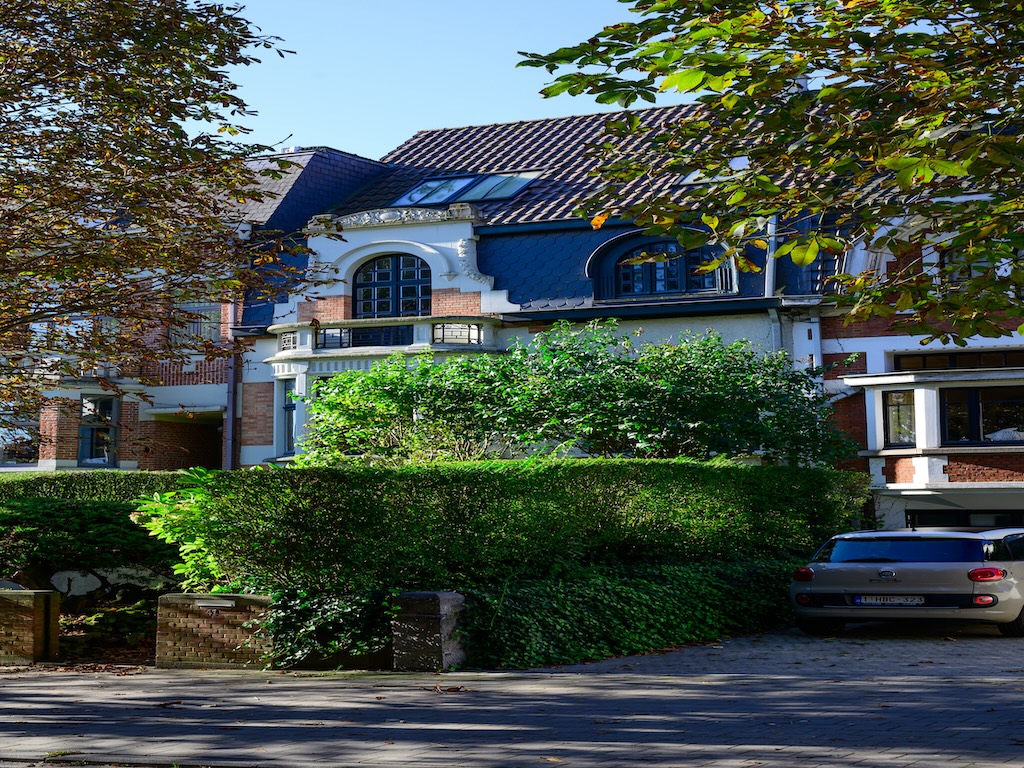 Maison moderne avec toit bleu, jardin paysager, balcon, allée pavée et vue dégagée.