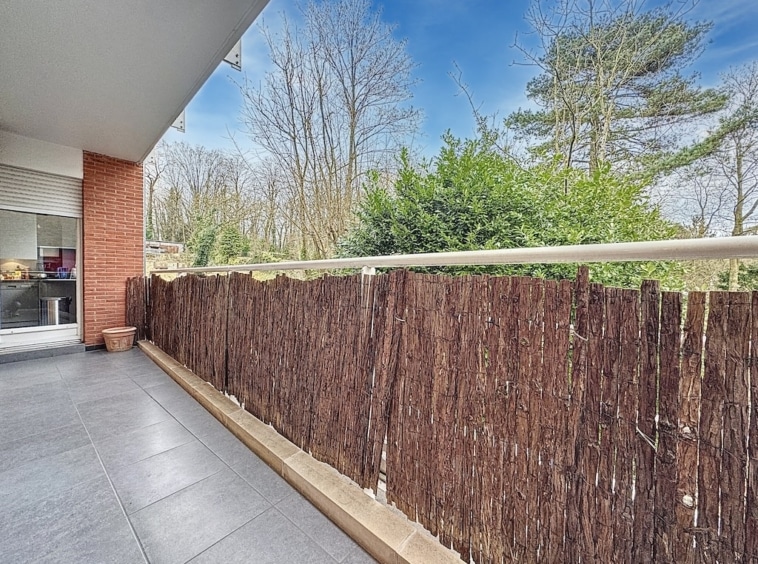 Balcon moderne avec vue sur nature verdoyante, clôture en branches et intérieur lumineux.