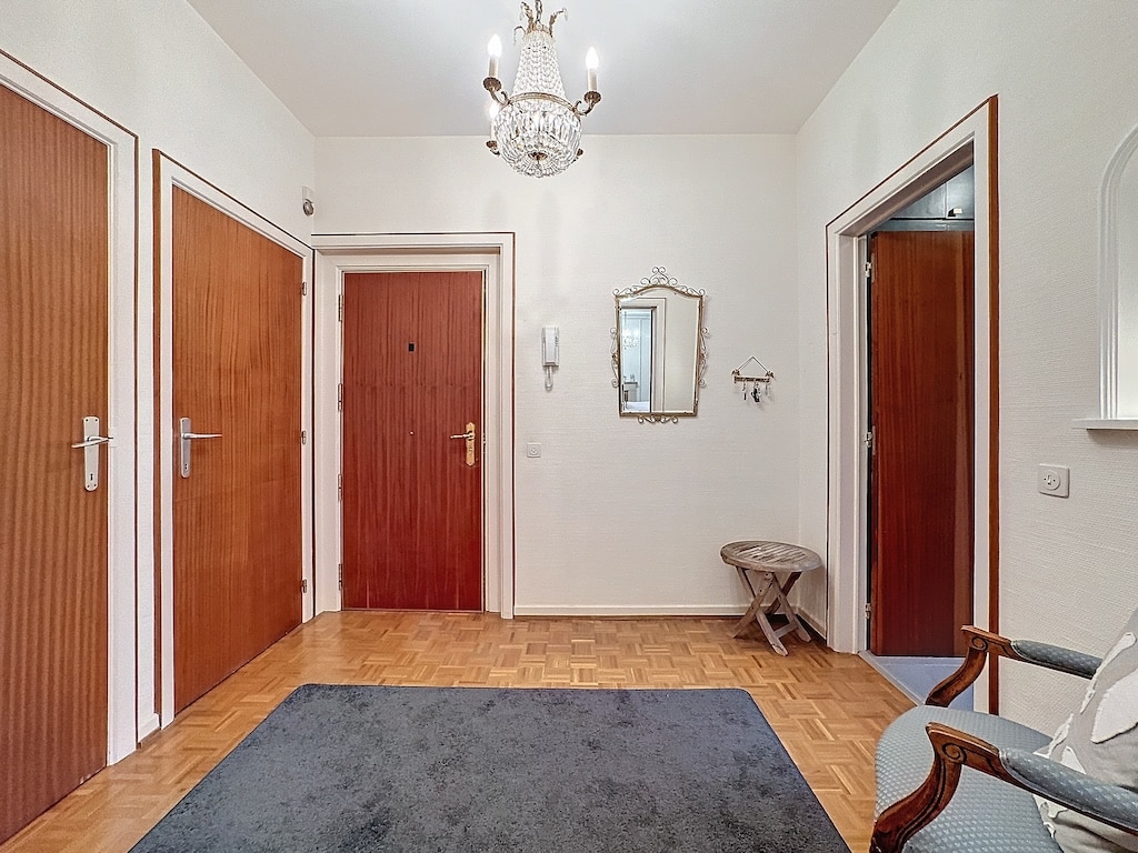 Entrée accueillante avec chandelier, miroir vintage, tapis gris, portes en bois, et décor élégant.