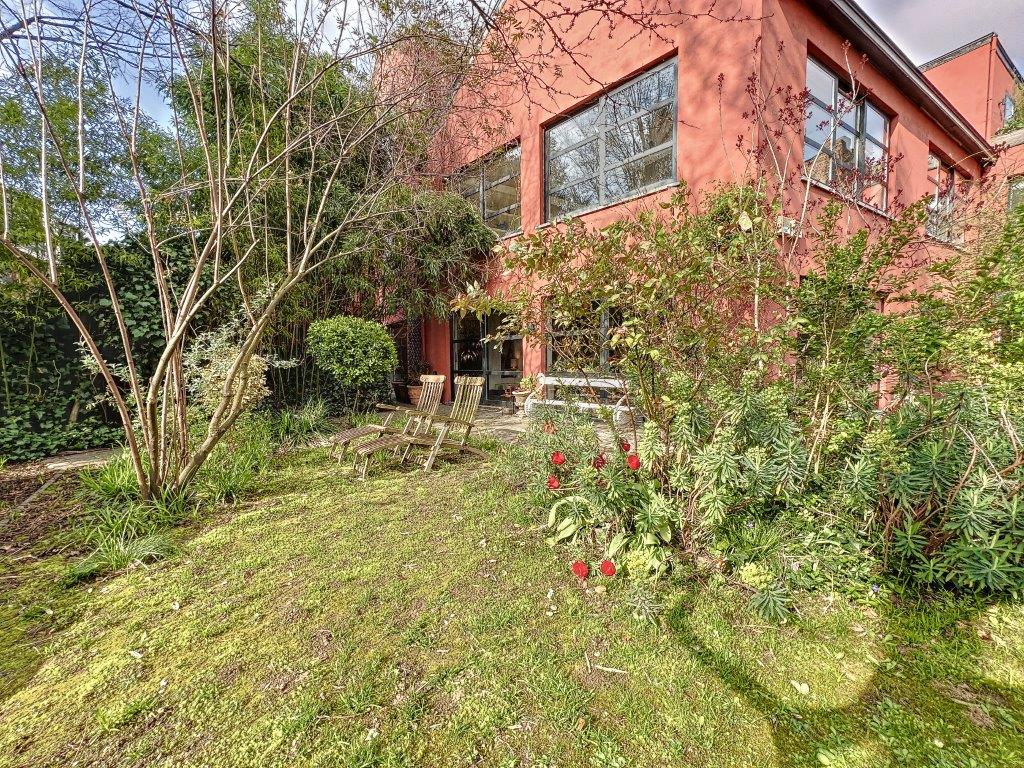 Jardin zen avec maison rouge moderne, fleurs colorées, chaises en bois et ambiance relaxante.