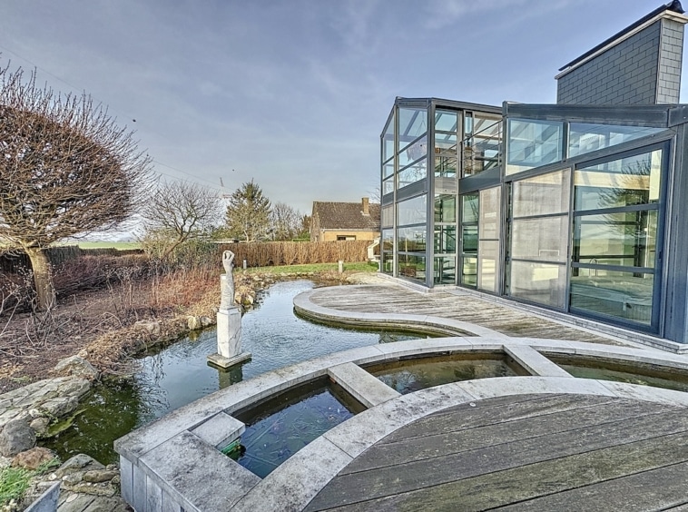 Maison moderne avec grande façade vitrée, bassin deau, chemin en bois et vue sur la nature.