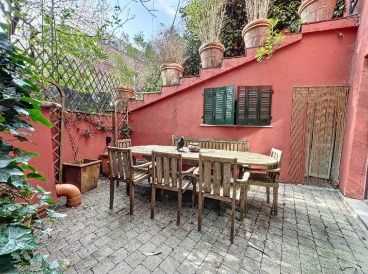 Patio ensoleillé avec mobilier en bois, mur rouge, plantes, ambiance chaleureuse et conviviale.