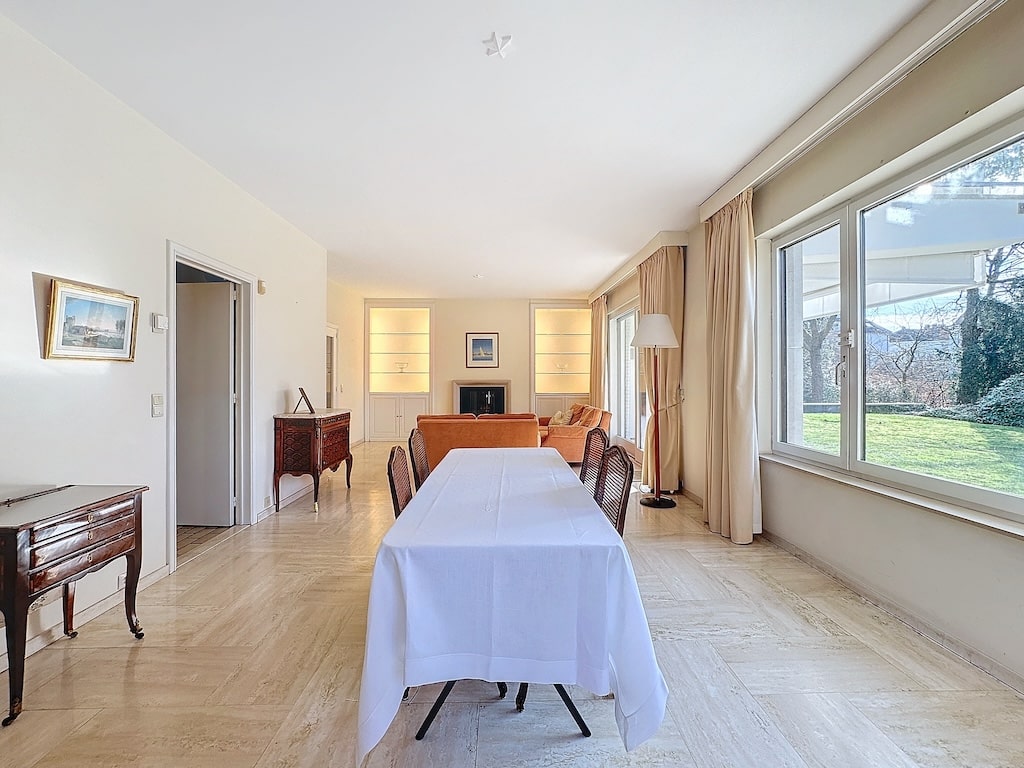 Salle à manger moderne avec grande table blanche, vue sur jardin verdoyant et canapé orange lumineux.