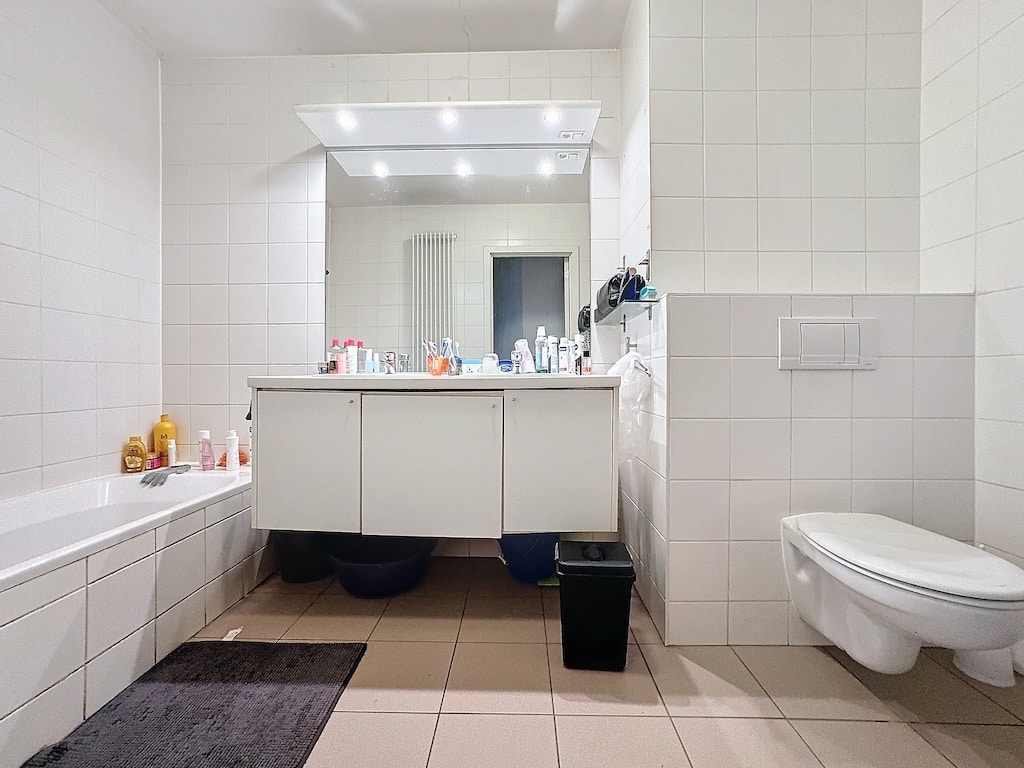 Salle de bain moderne blanche minimaliste avec baignoire, toilettes murales et grand miroir lumineux.