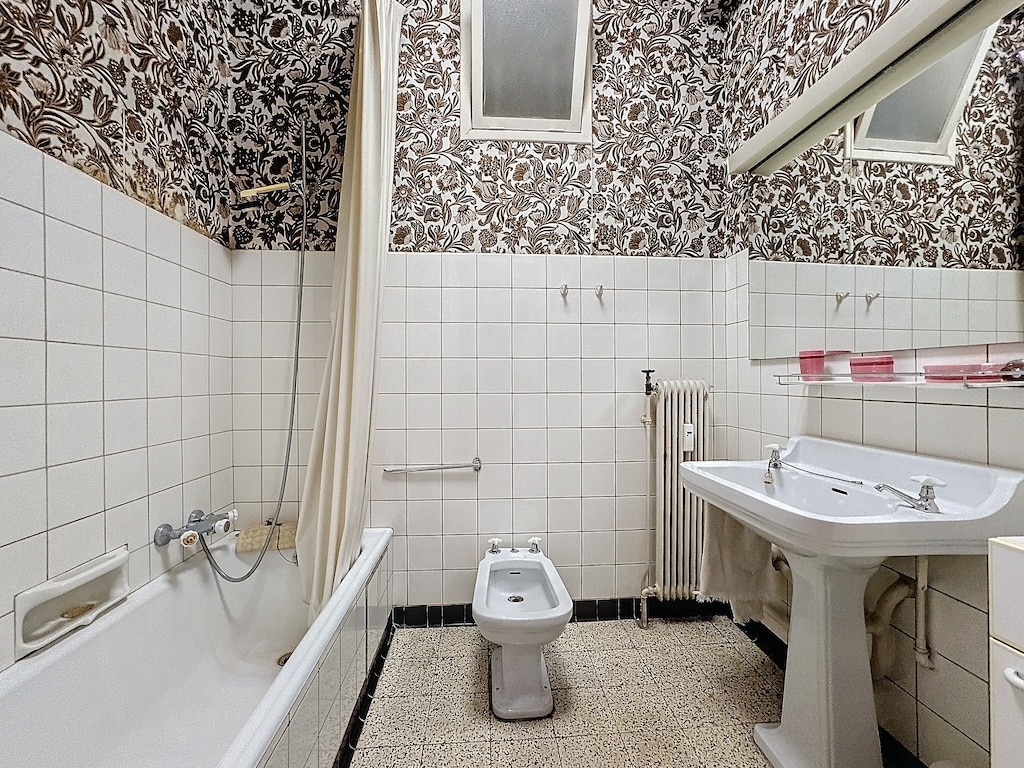 Salle de bain vintage charmante avec faïence blanche, papier floral sombre et sol en galets.
