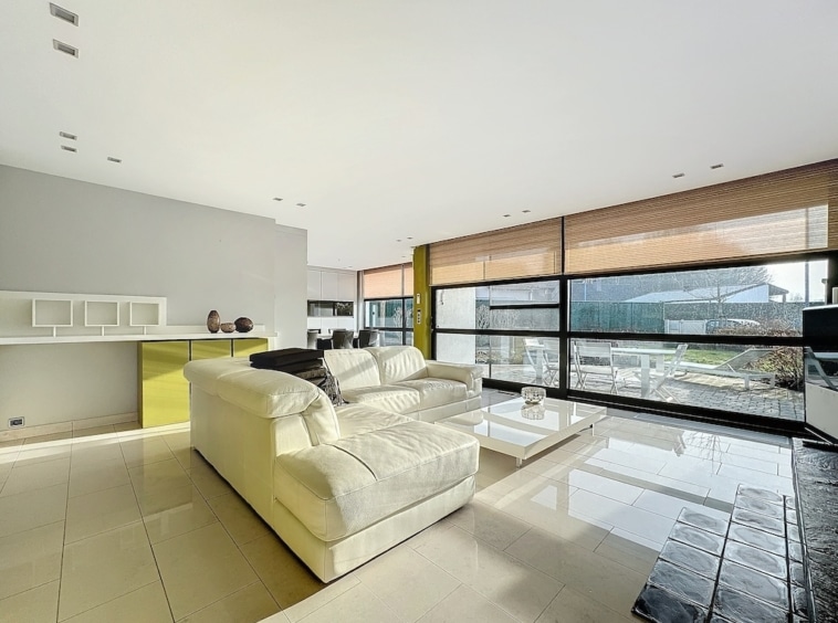 Salon moderne lumineux avec large baie vitrée, canapé blanc, bibliothèque blanche et mur jaune vivo.