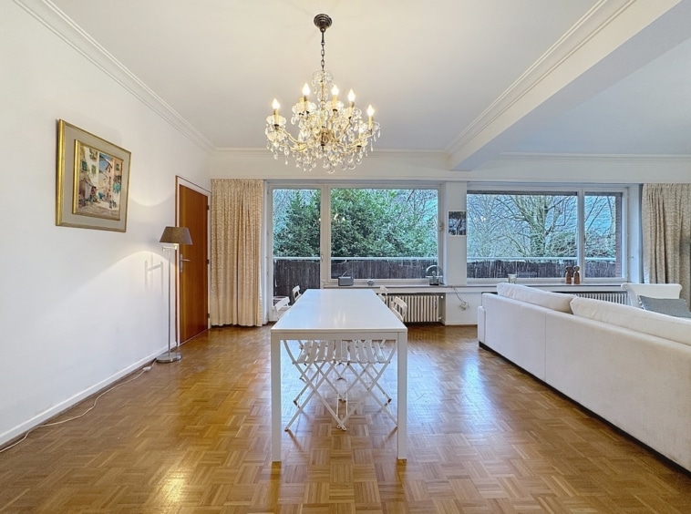 Salon lumineux avec chandelier en cristal, mobilier moderne et vue verdoyante.