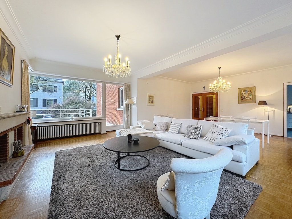 Salon chic et lumineux avec vue nature, canapé blanc, tableau, chandelier classique, tapis neutre.