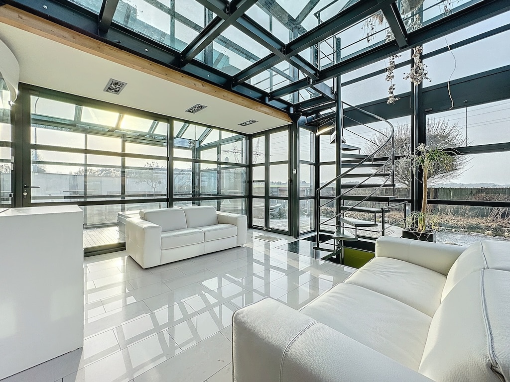 Salon moderne lumineux avec grandes fenêtres, mobilier blanc épuré, escalier en verre et plantes suspendues.