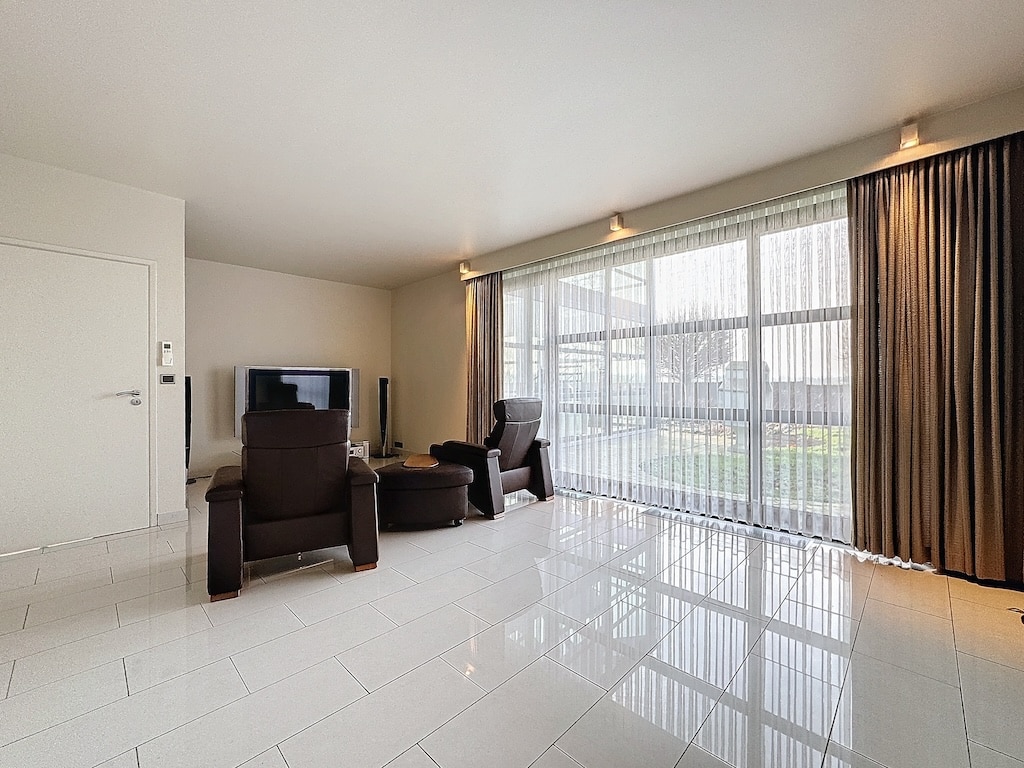 Salon moderne lumineux avec fauteuils, grandes fenêtres, décoration épurée et vue sur la verdure.