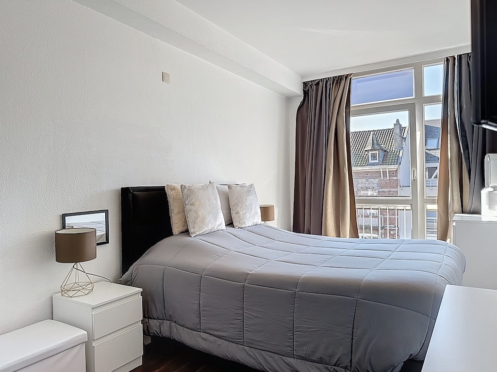 Chambre moderne, lumineuse et paisible avec lit gris, parquet foncé et lumière naturelle douce.