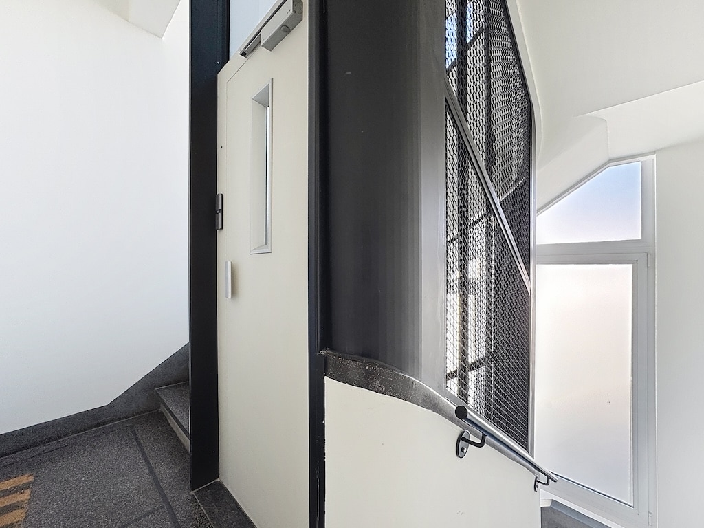 Escalier moderne en coin avec mur blanc, rampe métallique et porte lumineuse.