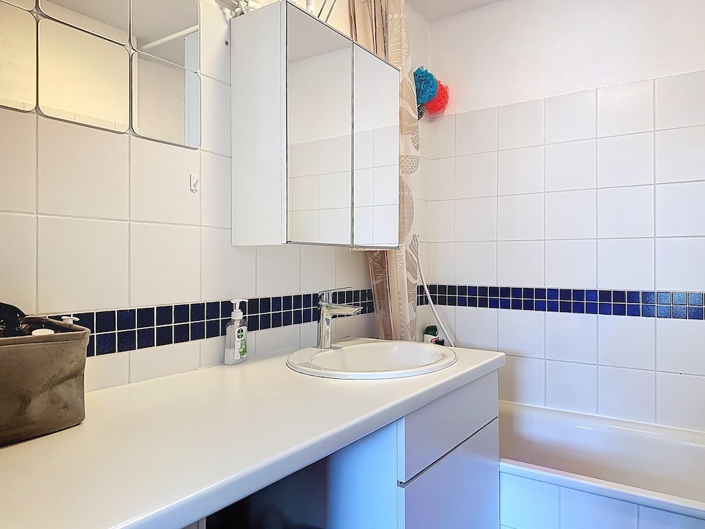 Salle de bain lumineuse minimaliste, touches de bleu, miroir, baignoire, accessoires colorés.