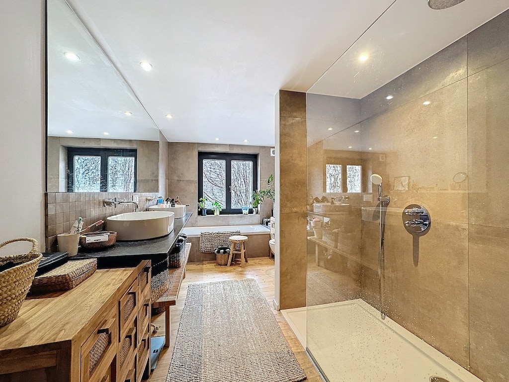 Salle de bain moderne avec baignoire, douche en verre, meuble en bois, plantes et lumière douce.