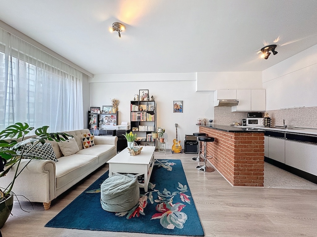 Salon lumineux moderne avec canapé blanc, tapis coloré et cuisine en briques rustique.