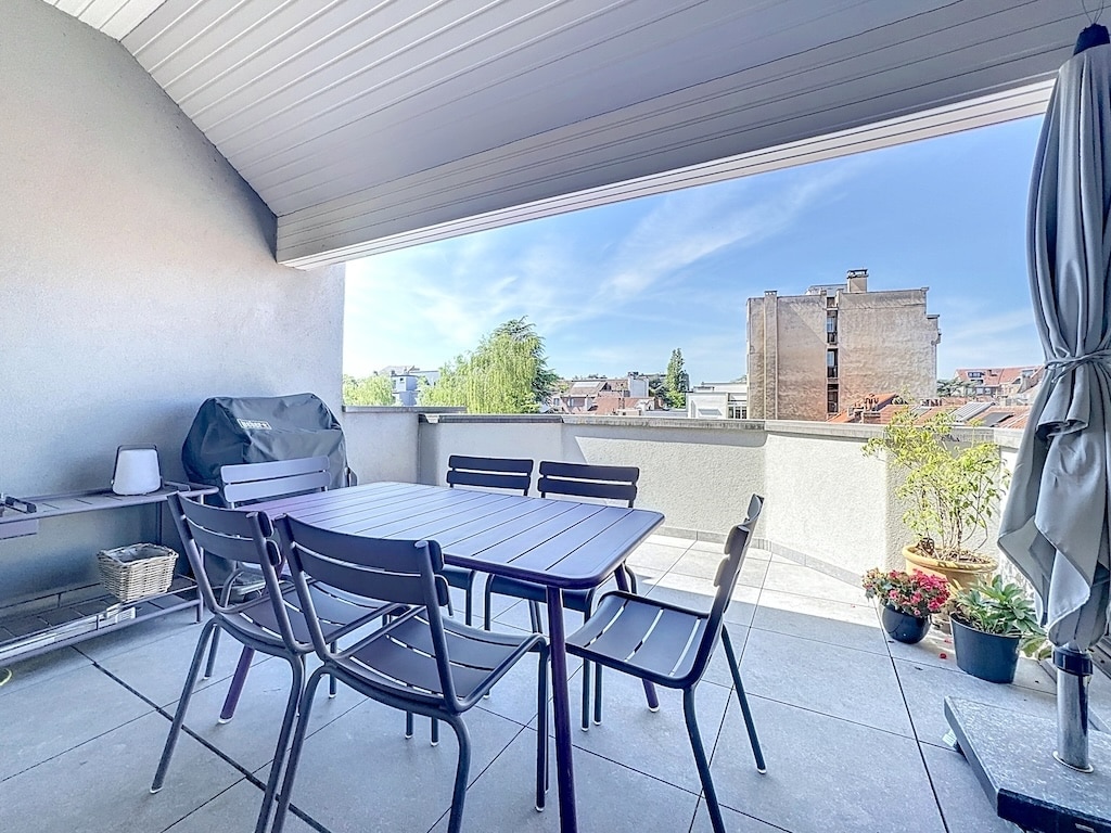 Balcon contemporain avec mobilier en métal, barbecue, plantes, vue urbaine verdoyante et ambiance chaleureuse.