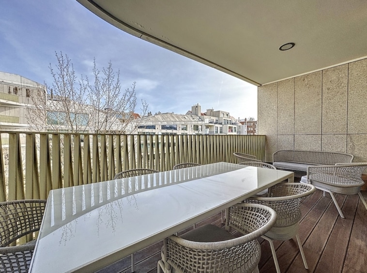 Balcon contemporain avec table en verre, chaises en osier, vue sur arbres nus et ville.