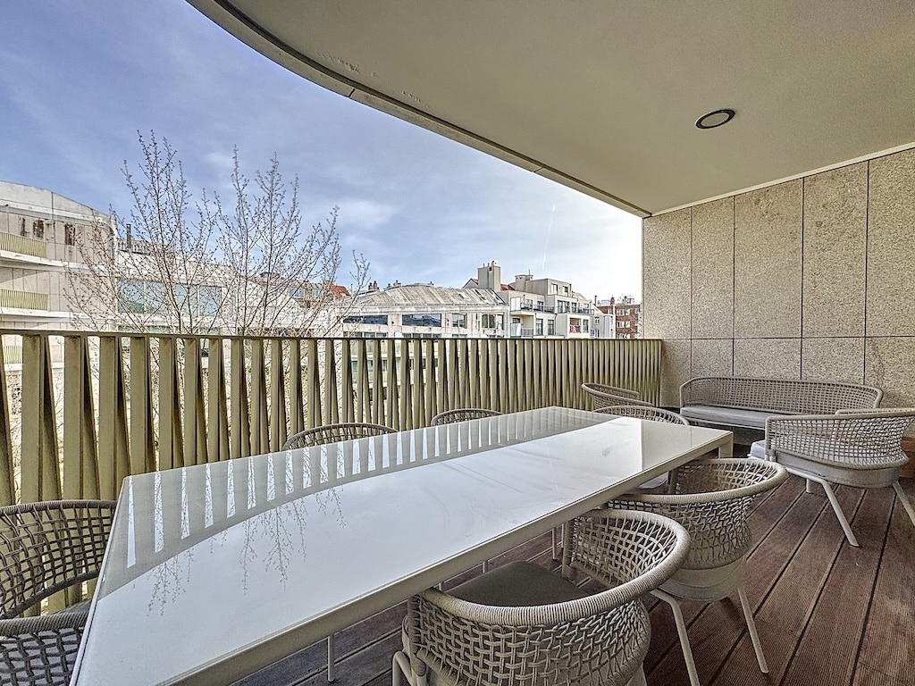 Balcon contemporain avec table en verre, chaises en osier, vue sur arbres nus et ville.