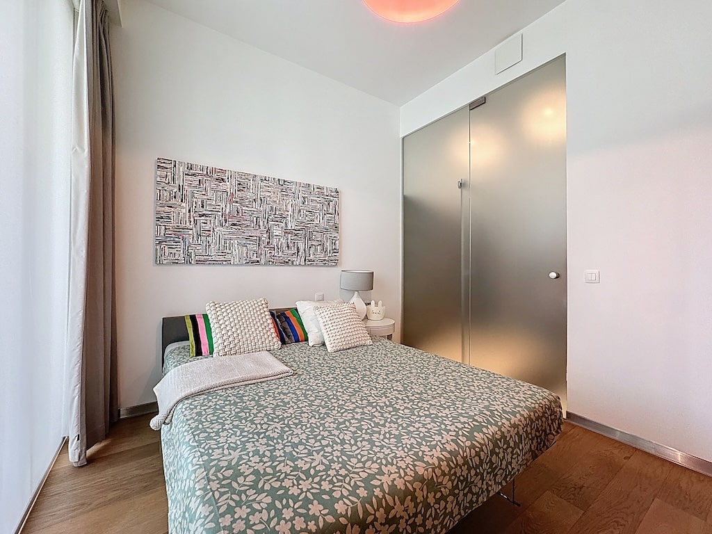 Chambre minimaliste moderne avec lit coloré, art abstrait, porte vitrée et lumière naturelle abondante.