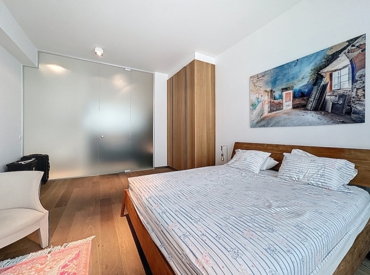 Chambre minimaliste avec grand lit en bois, œuvre dart colorée, fauteuil, tapis et séparation en verre.