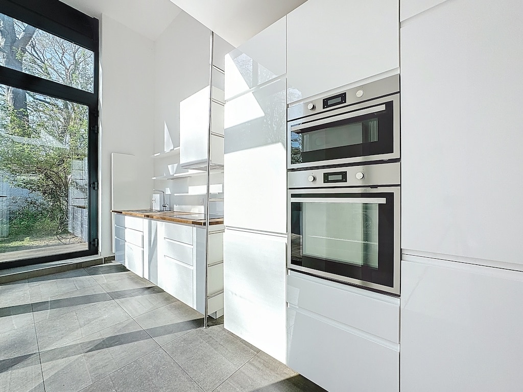 Cuisine moderne lumineuse avec armoires blanches, grands fours en acier inoxydable, sol gris épuré.