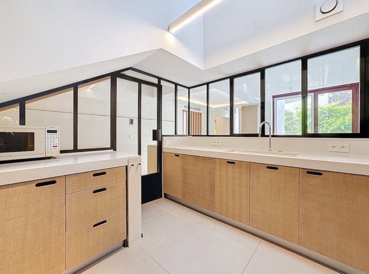 Cuisine moderne lumineuse avec grandes fenêtres, armoires en bois clair et vue sur la verdure.