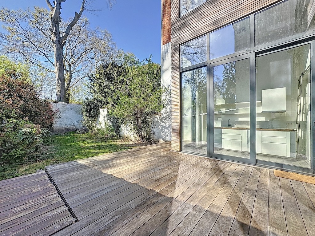 Espace extérieur moderne avec terrasse en bois, jardin verdoyant et vue intérieure lumineuse.