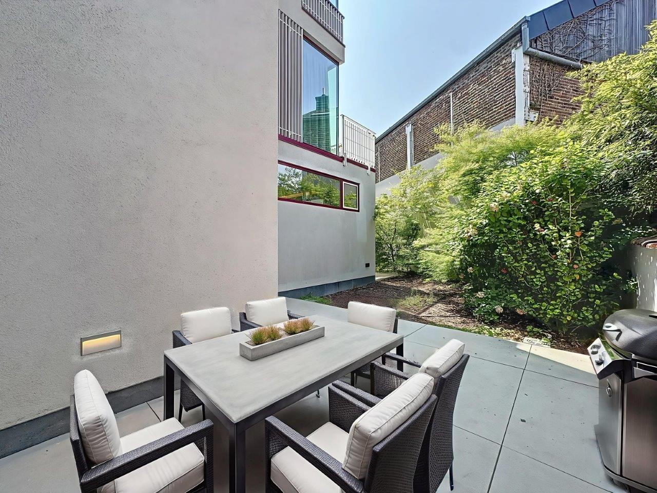 Espace repas extérieur moderne avec mobilier élégant, végétation luxuriante et barbecue en acier inoxydable.