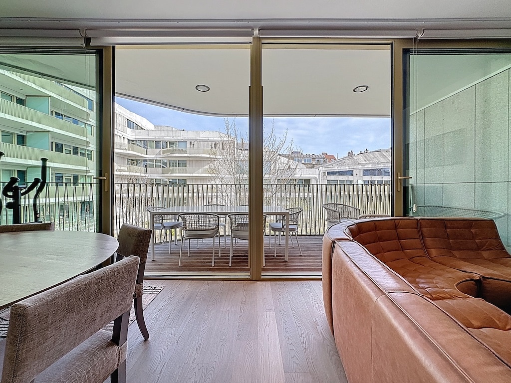 Salon moderne lumineux avec balcon urbain, canapé en cuir, espace convivial et vue sur la ville.