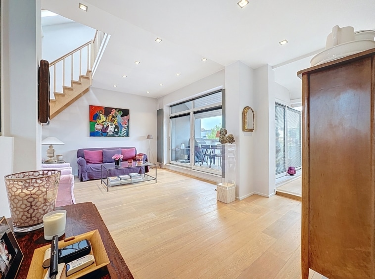 Intérieur moderne lumineux avec canapé violet, escalier en bois, grandes fenêtres et décoration chaleureuse.