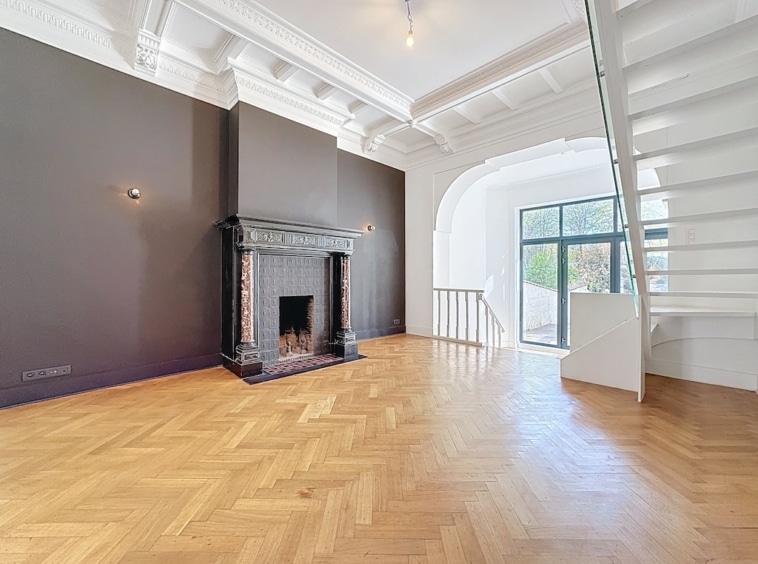 Salon élégant avec parquet en chevrons, cheminée classique, grandes fenêtres et escalier moderne blanc.