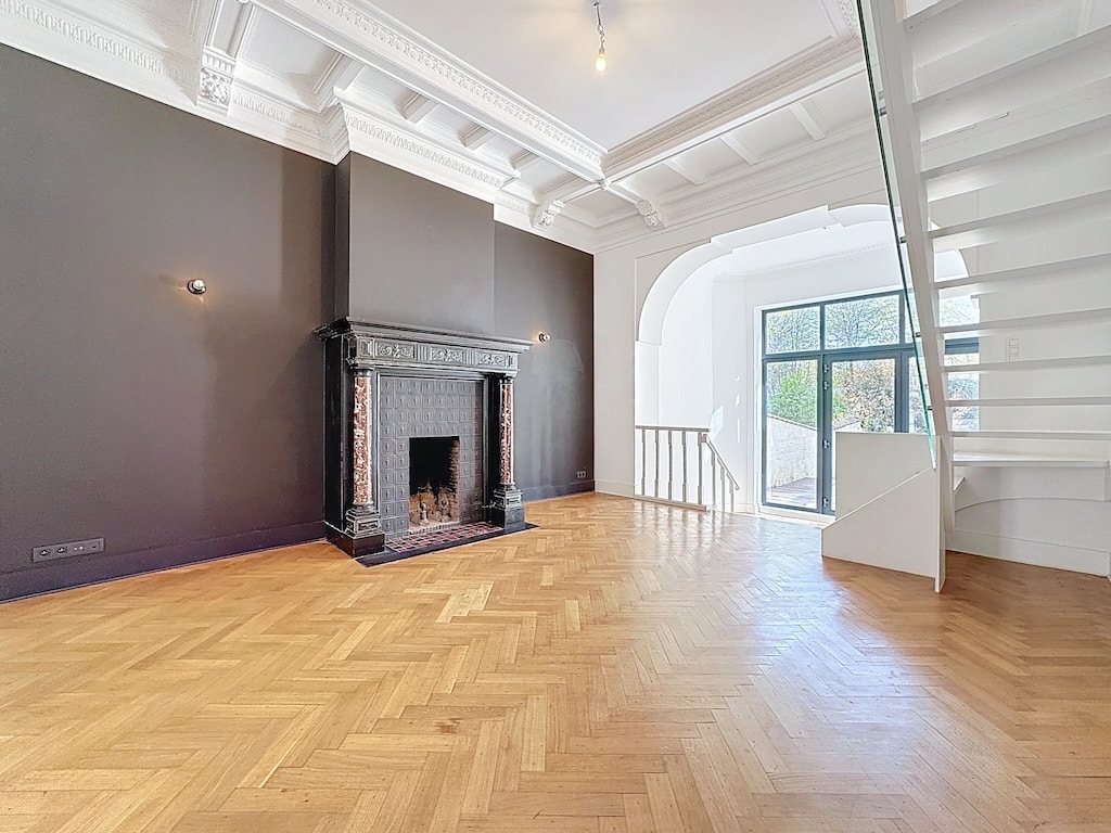 Salon élégant avec parquet en chevrons, cheminée classique, grandes fenêtres et escalier moderne blanc.