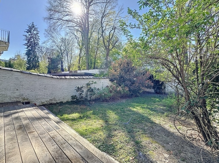Jardin paisible ensoleillé avec terrasse en bois, mur blanc et végétation luxuriante.