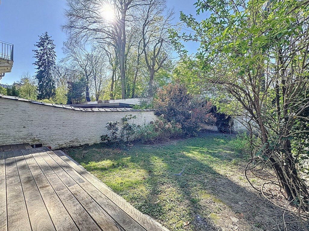 Jardin paisible ensoleillé avec terrasse en bois, mur blanc et végétation luxuriante.