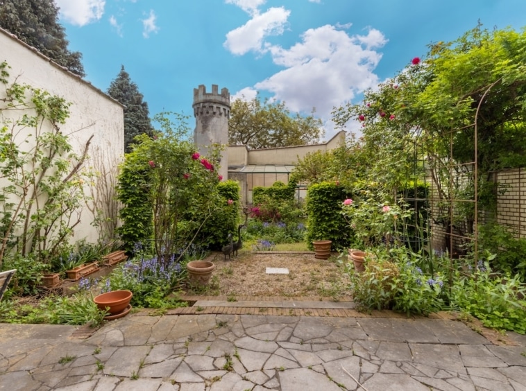 Jardin féerique avec tour, chemin pavé et fleurs colorées sous un ciel bleu.