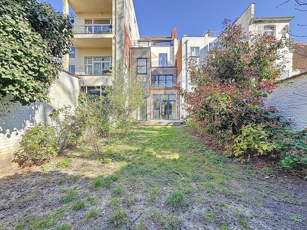 Jardin moderne paisible avec végétation luxuriante et vue sur résidence contemporaine ensoleillée.