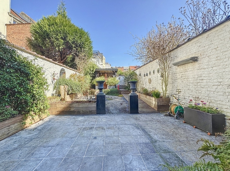 Jardin paisible avec chemin en pierre, urnes élégantes, murs blancs, et plantes grimpantes.