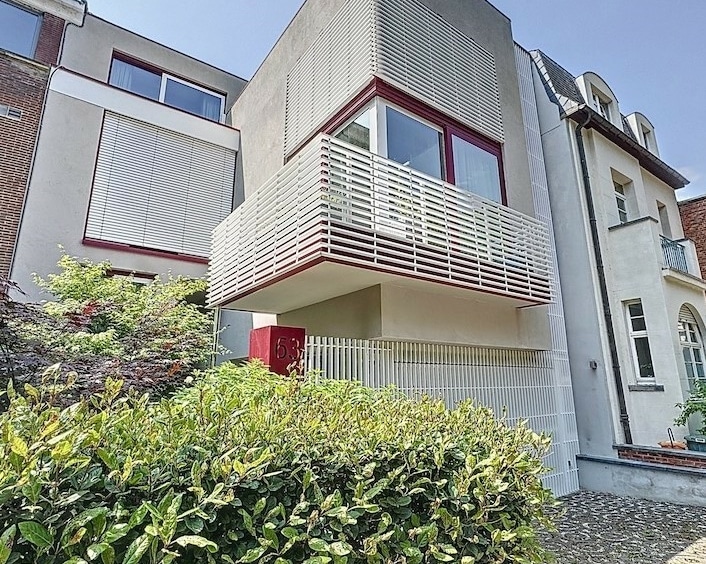 Maison moderne minimaliste avec balcon en verre et porte rouge, contrastant avec bâtiment traditionnel.
