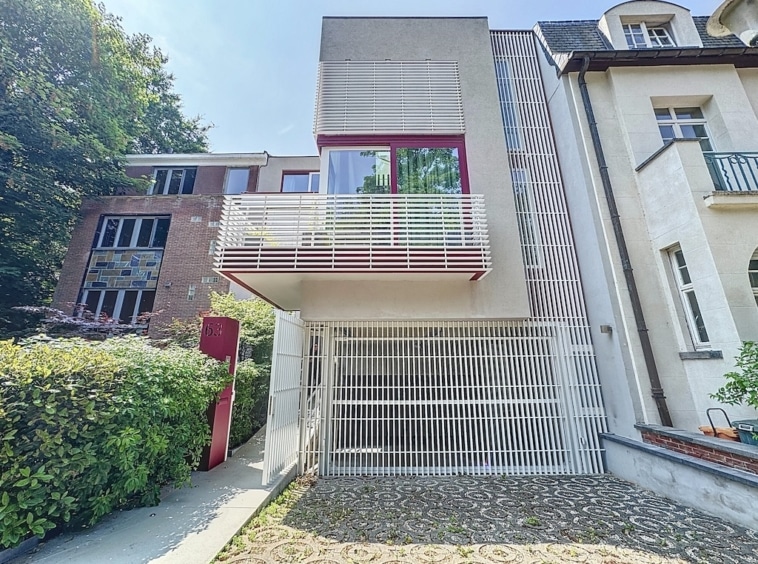 Habitation moderne avec balcon en verre, façade en béton, bois et touches de rouge, entourée de verdure.