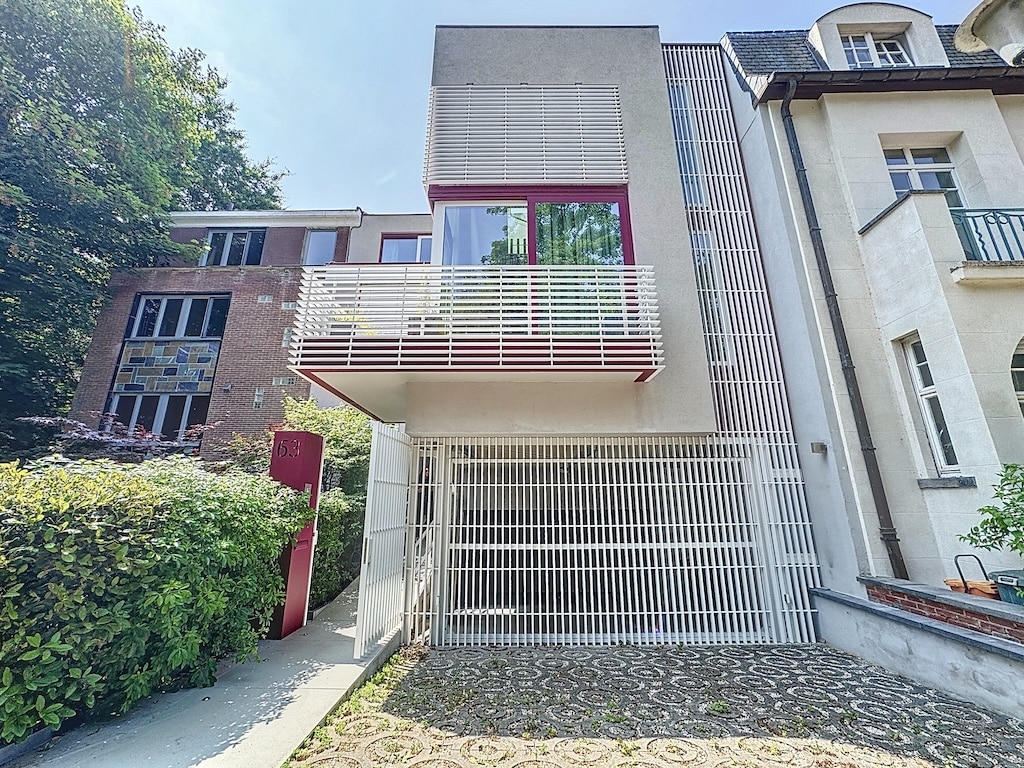 Habitation moderne avec balcon en verre, façade en béton, bois et touches de rouge, entourée de verdure.