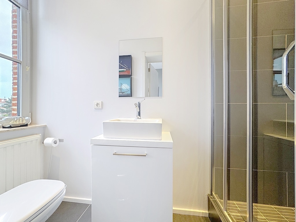Salle de bain moderne, lumineuse, avec douche en verre, WC blanc et mobilier épuré.