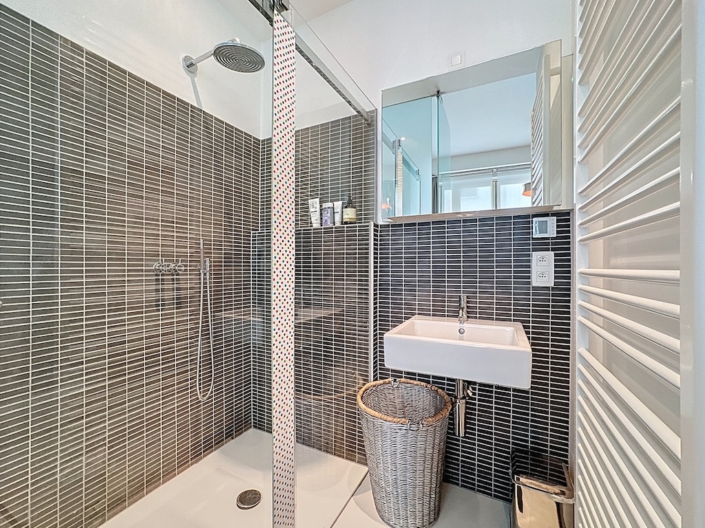 Salle de bain moderne avec douche en verre, murs sombres, évier mural et sèche-serviettes chaleureux.