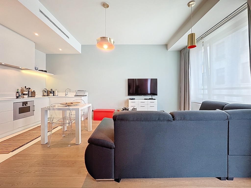 Salon moderne lumineux avec canapé noir, cuisine ouverte blanche, mobilier épuré et ambiance chaleureuse.